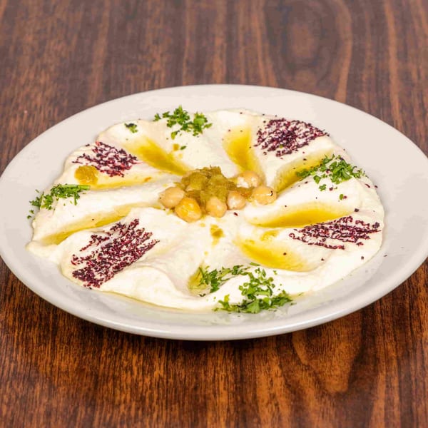 A white plate of hummus garnished with chickpeas, green relish, olive oil, sumac, and parsley.