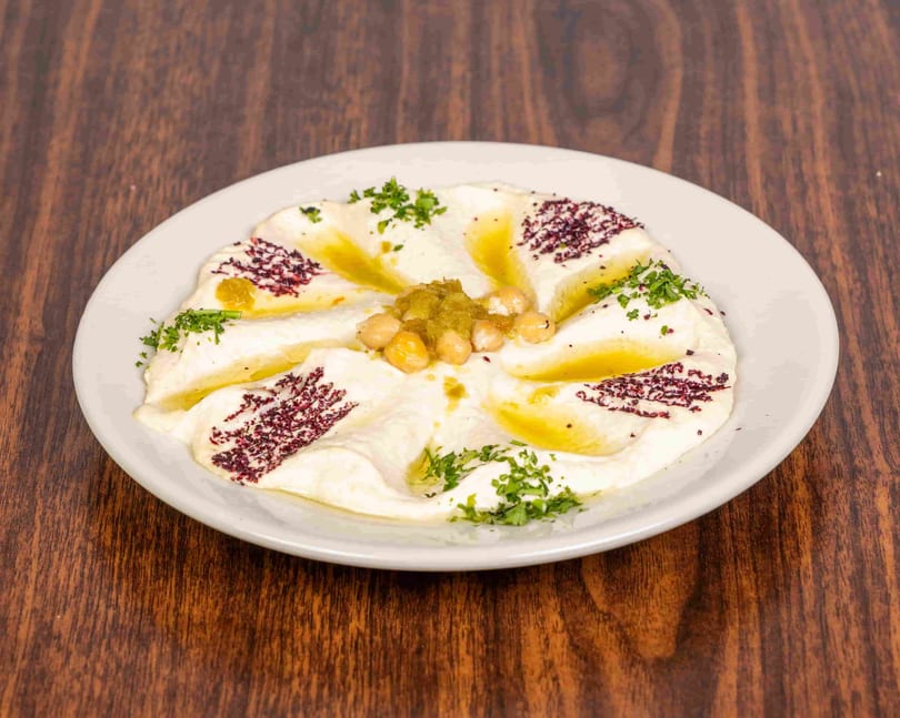A white plate of hummus garnished with chickpeas, green relish, olive oil, sumac, and parsley.