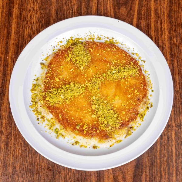Golden kunafa dessert topped with crushed pistachios on a white plate, on a wooden table.