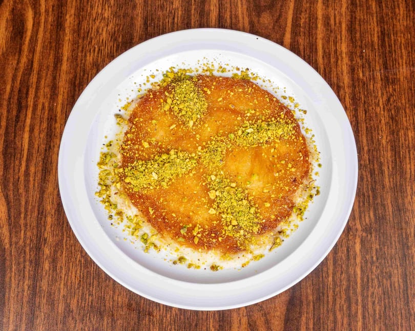Golden kunafa dessert topped with crushed pistachios on a white plate, on a wooden table.