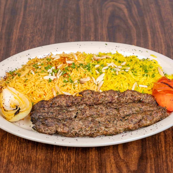 A plate of kofta kebab with yellow rice, grilled onion, and tomato on a wooden table.