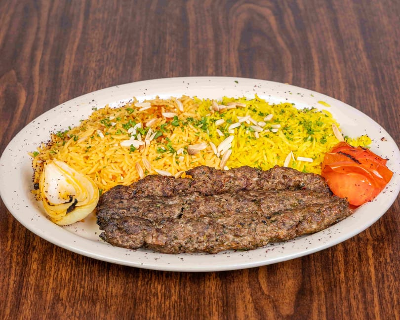 A plate of kofta kebab with yellow rice, grilled onion, and tomato on a wooden table.