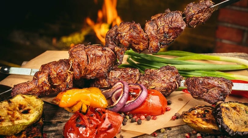 Grilled meat skewers with roasted vegetables like bell peppers, onions, and zucchini, against a fiery background.