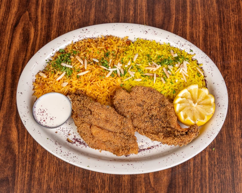 Two pieces of fried fish with yellow and orange rice, lemon, and a side of white sauce.