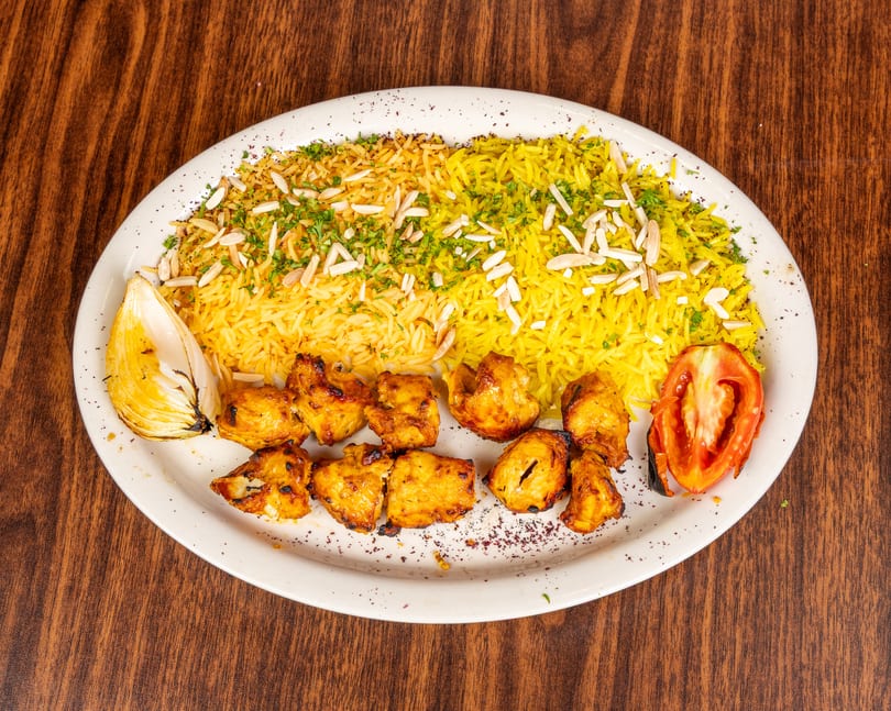 Chicken shish tawook with yellow rice, almonds, parsley, grilled onion, and tomato on a white plate.