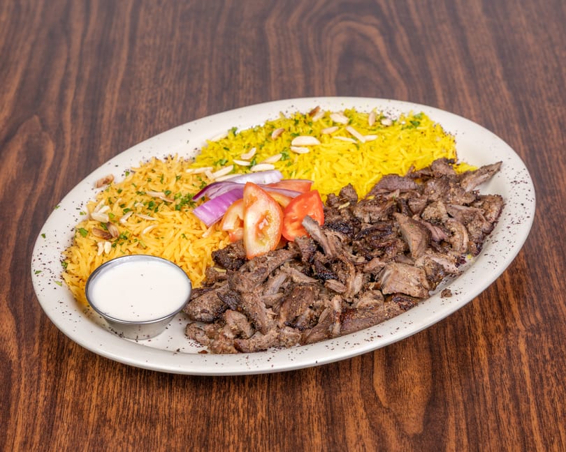 Shawarma plate with seasoned meat, yellow rice, sliced tomatoes, red onions, and a side of white sauce.