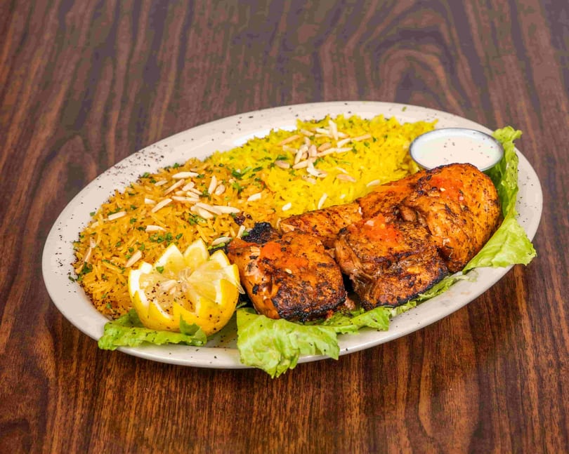 Grilled chicken with yellow rice, garnished with almonds, parsley, lemon, and a side of sauce.