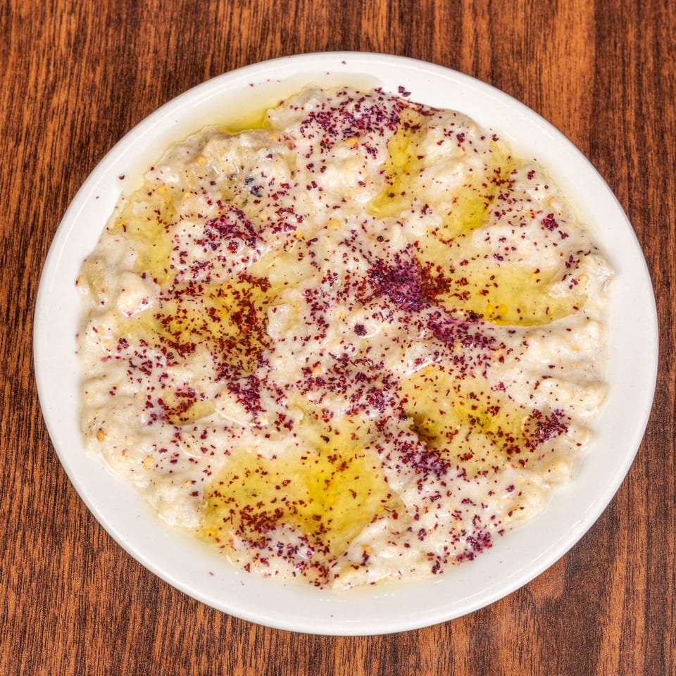 A white plate of creamy baba ghanoush topped with olive oil and red spice on a wooden table.