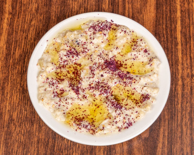 A white plate of creamy baba ghanoush topped with olive oil and red spice on a wooden table.