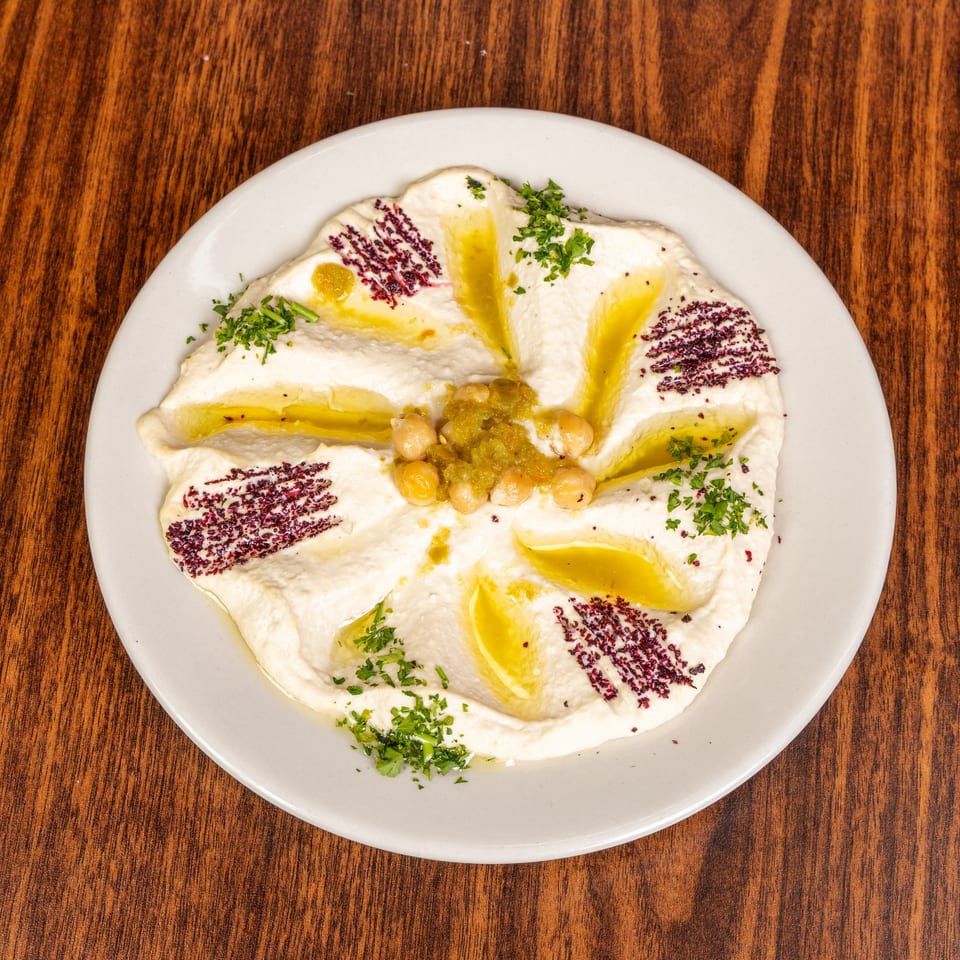 Creamy hummus drizzled with olive oil, garnished with chickpeas, green relish, sumac, and parsley.