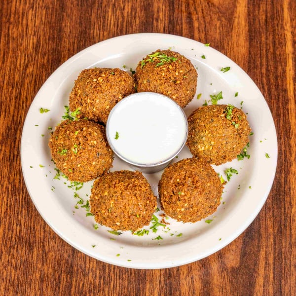 Six golden-brown falafel balls with sesame seeds and parsley, surrounding a white dipping sauce on a plate.