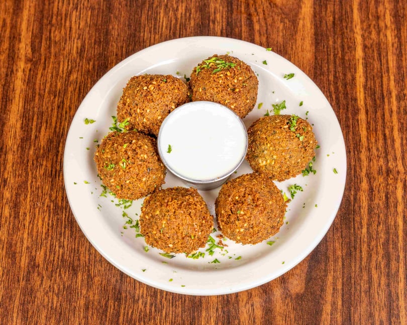Six golden-brown falafel balls with sesame seeds and parsley, surrounding a white dipping sauce on a plate.