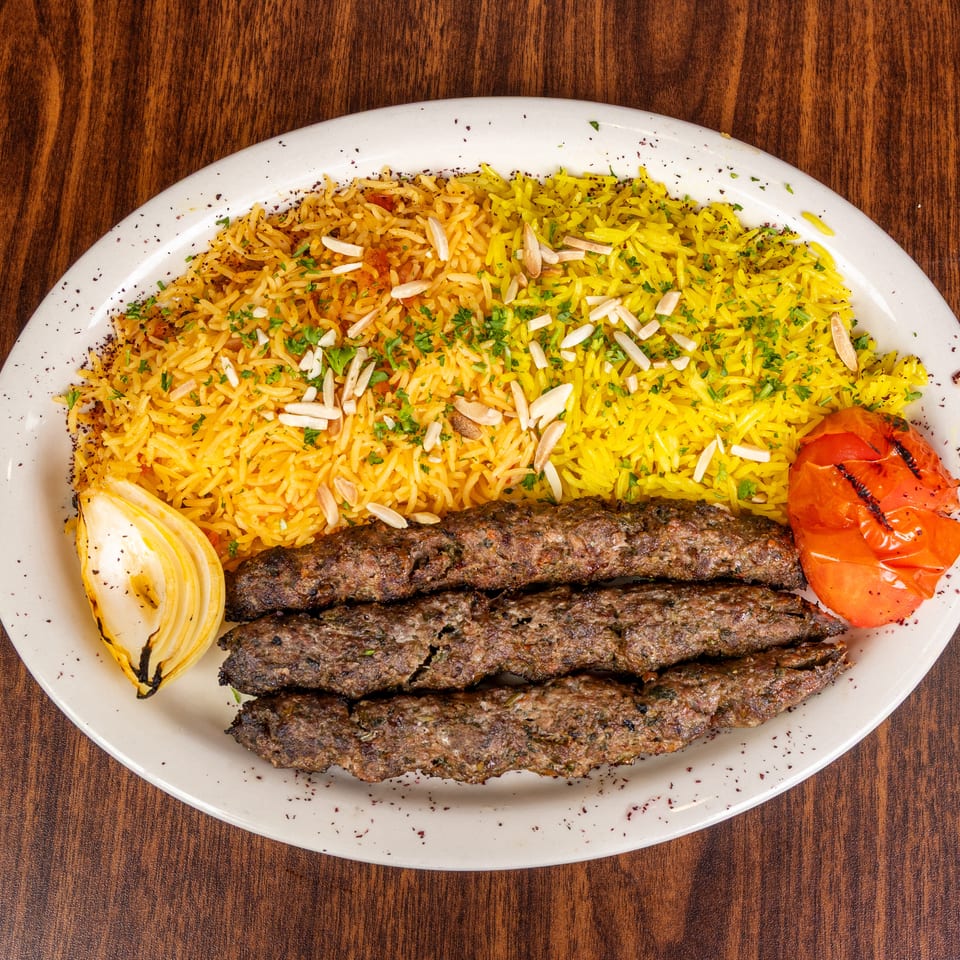 Three grilled meat skewers with yellow rice, toasted almonds, grilled onion, and tomato on a white plate.