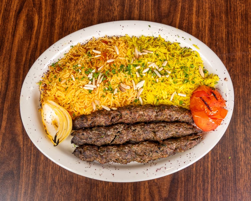 Three grilled meat skewers with yellow rice, toasted almonds, grilled onion, and tomato on a white plate.