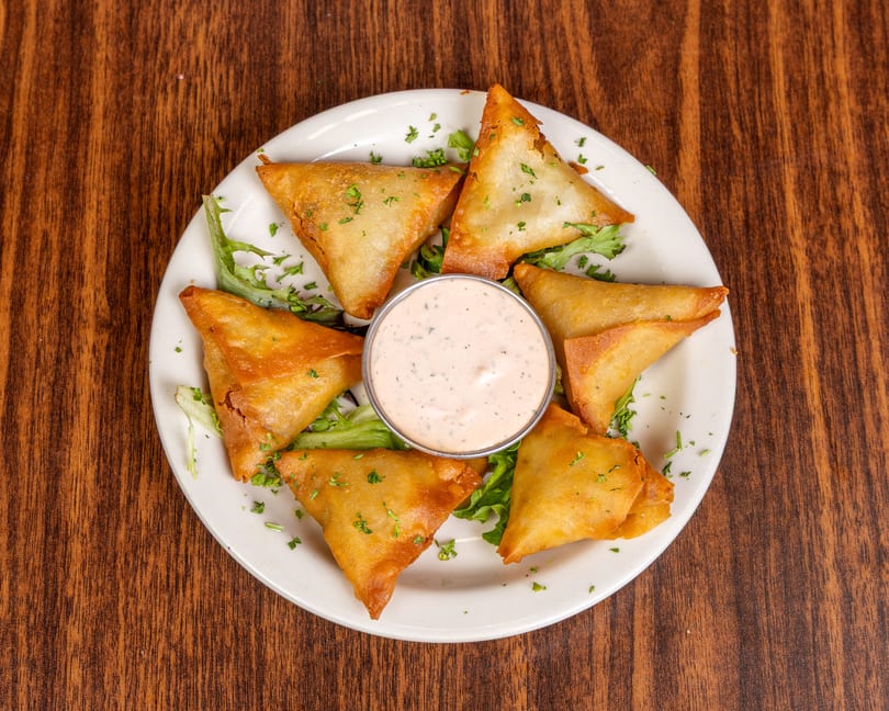 Golden-brown samosas arranged around a small bowl of dipping sauce on a white plate with lettuce.