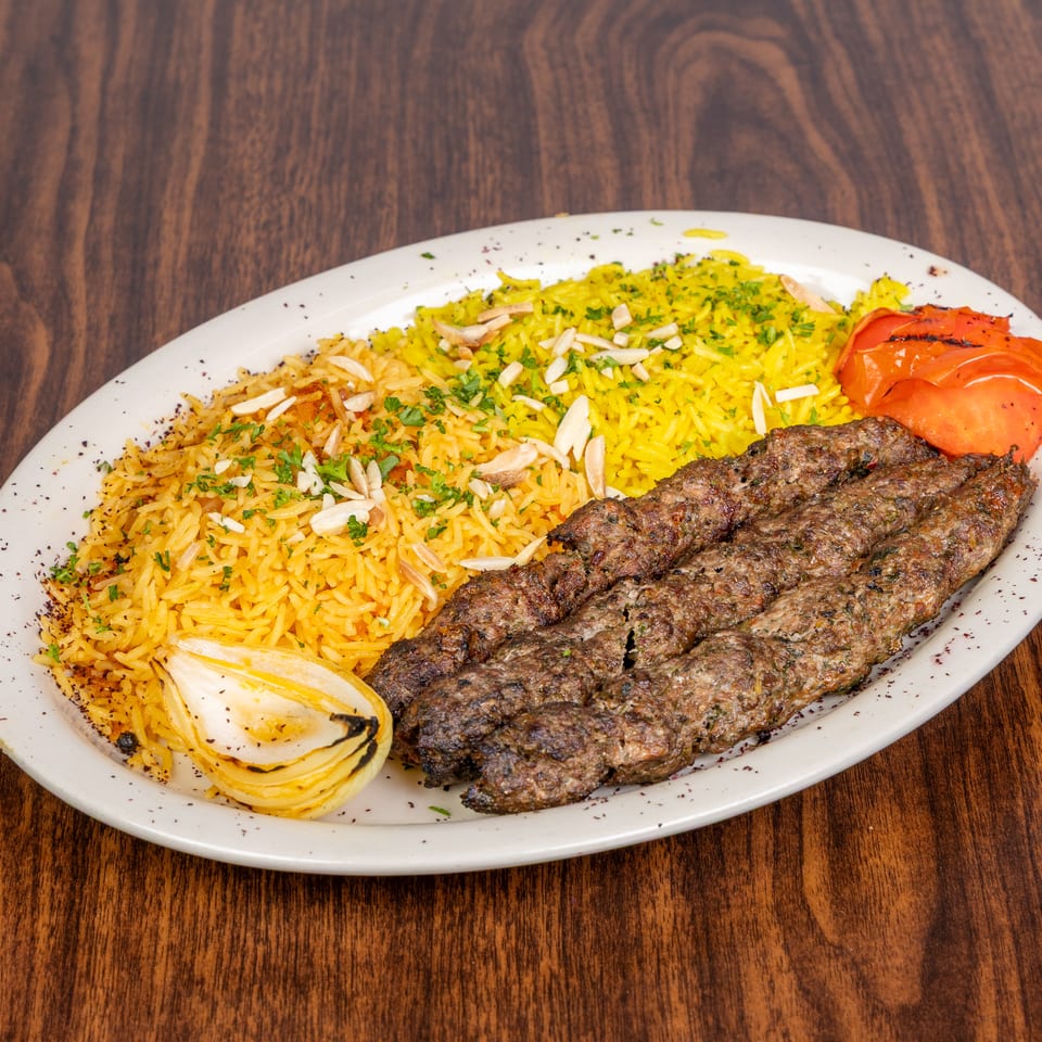 Three grilled meat skewers with yellow rice, roasted tomato, and onion on a white plate.