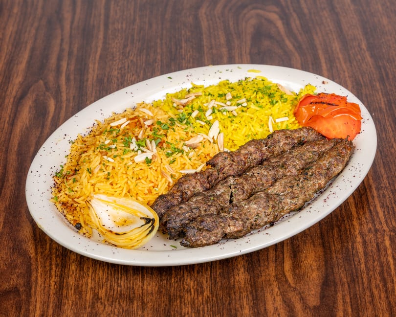 Three grilled meat skewers with yellow rice, roasted tomato, and onion on a white plate.