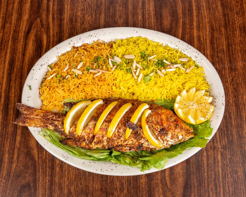 Whole grilled fish with lemon slices, served with yellow rice and lettuce on a white plate.