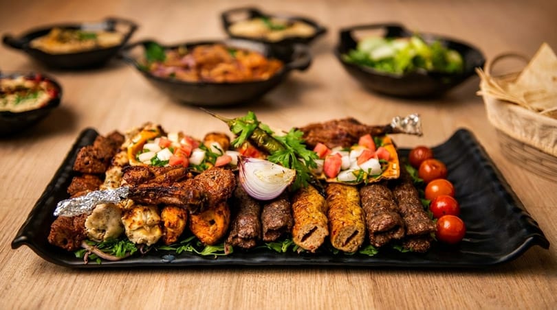 A large platter of grilled meats, vegetables, and sides on a wooden table.