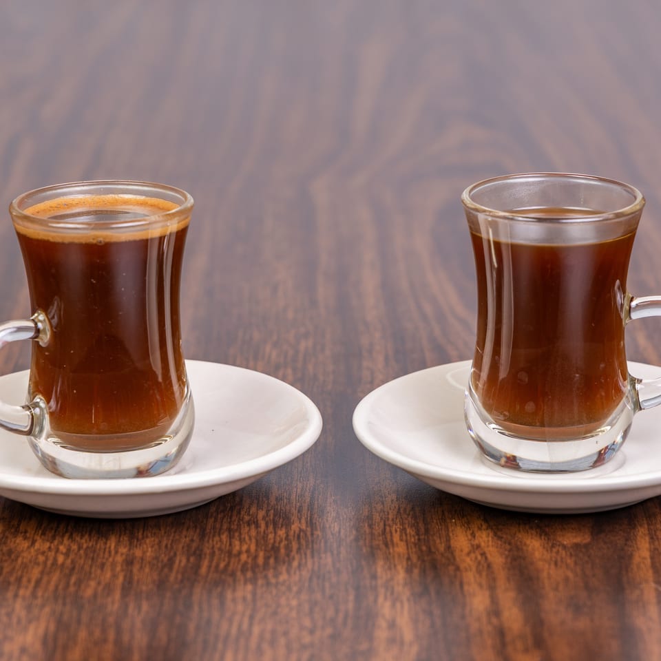 Two small glass cups of dark coffee with white saucers on a wooden table.