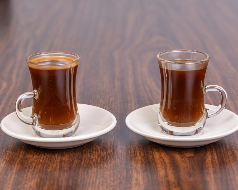 Two small glass cups of dark coffee with white saucers on a wooden table.